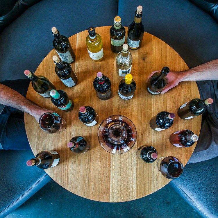 Two men sitting across from each other at a small round wooden table with a bunch of bottles and glasses of wine. Today we recommend 14 bottles of chilled red wine to drink this summer.