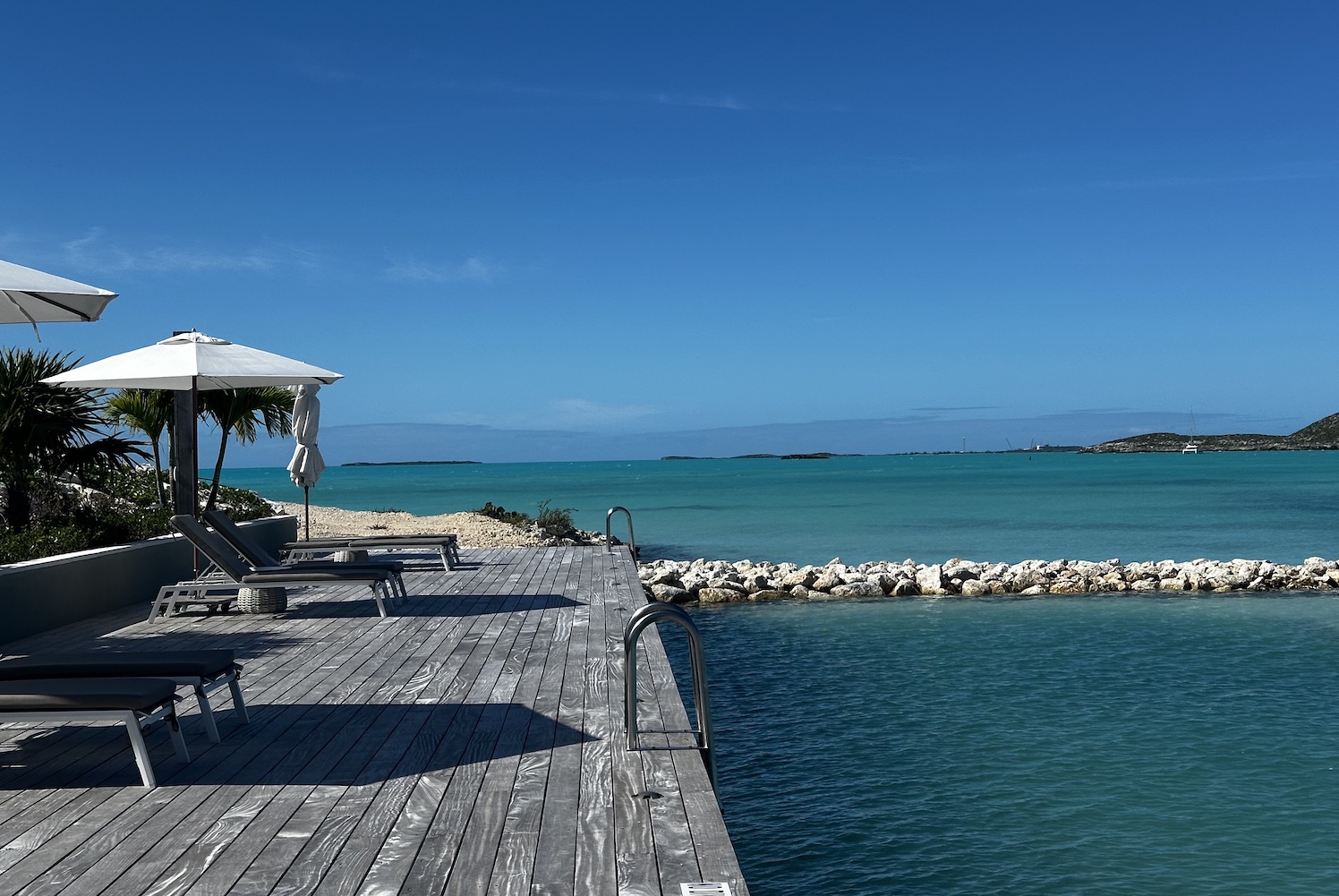 an ocean pool with blue skies and a grey wooden deck with chairs and white umbrellas