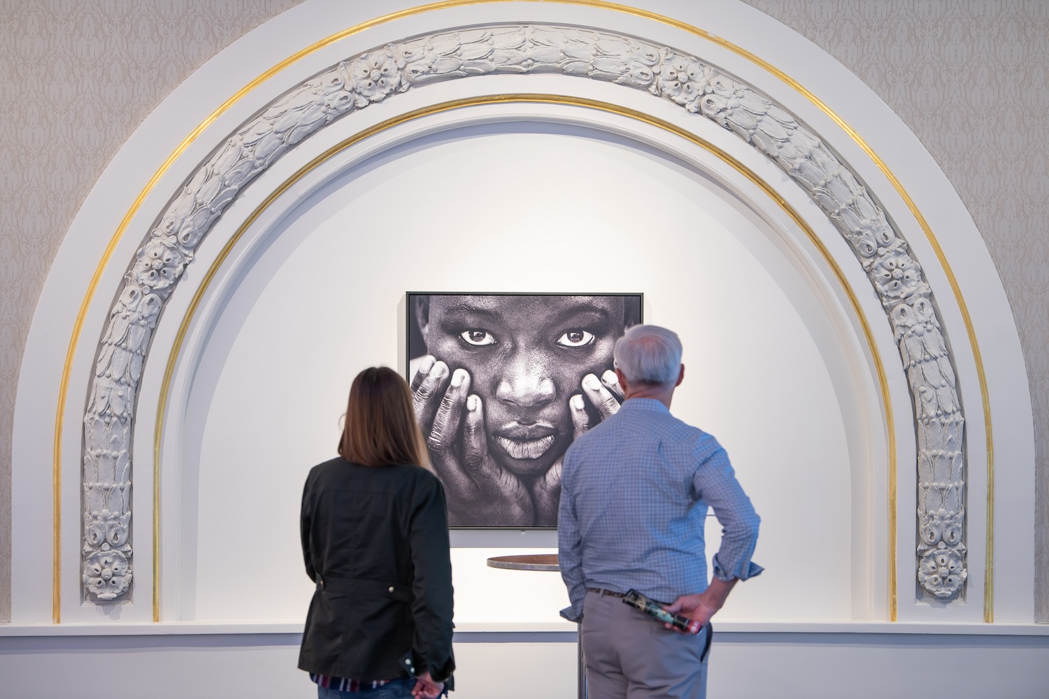 man and woman looking at art of a woman holding her face, looking directly forward, under a white arch