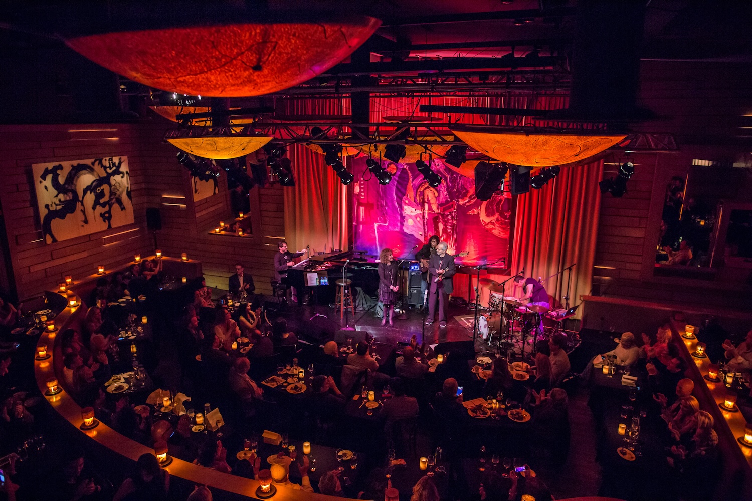 jazz group performing on stage, people sitting watching, red curtains, lights on stage, candles on tables