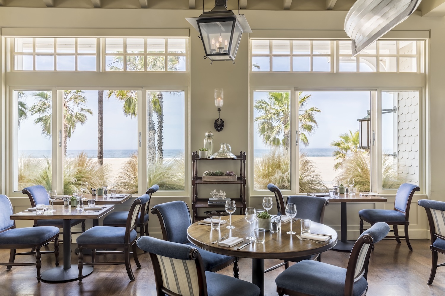 tables with glassware, blue chairs, windows facing beach and palm trees