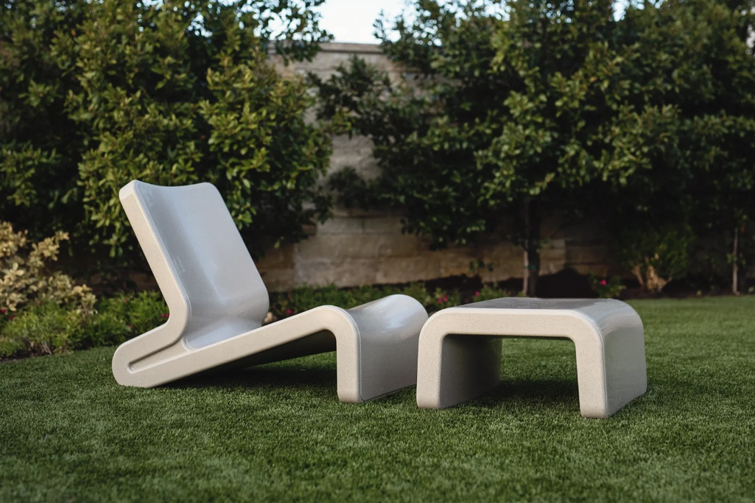 A grey lounge chair and ottoman sitting on grass in a backyard with a wall behind them