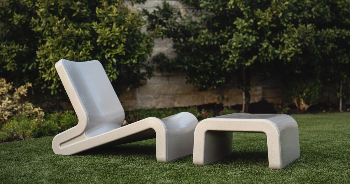 A grey lounge chair and ottoman sitting on grass in a backyard with a wall behind them