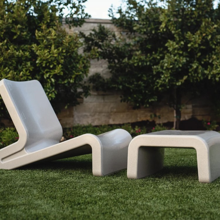 A grey lounge chair and ottoman sitting on grass in a backyard with a wall behind them