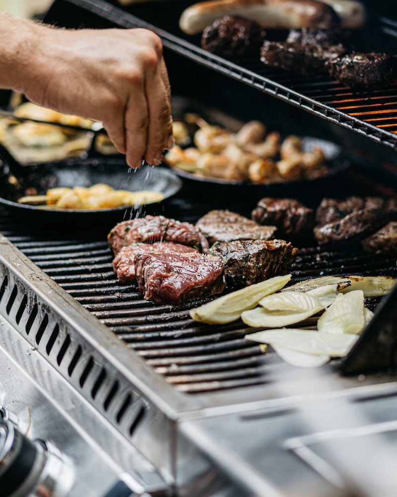 Human hand adds salt to the steaks on the grill