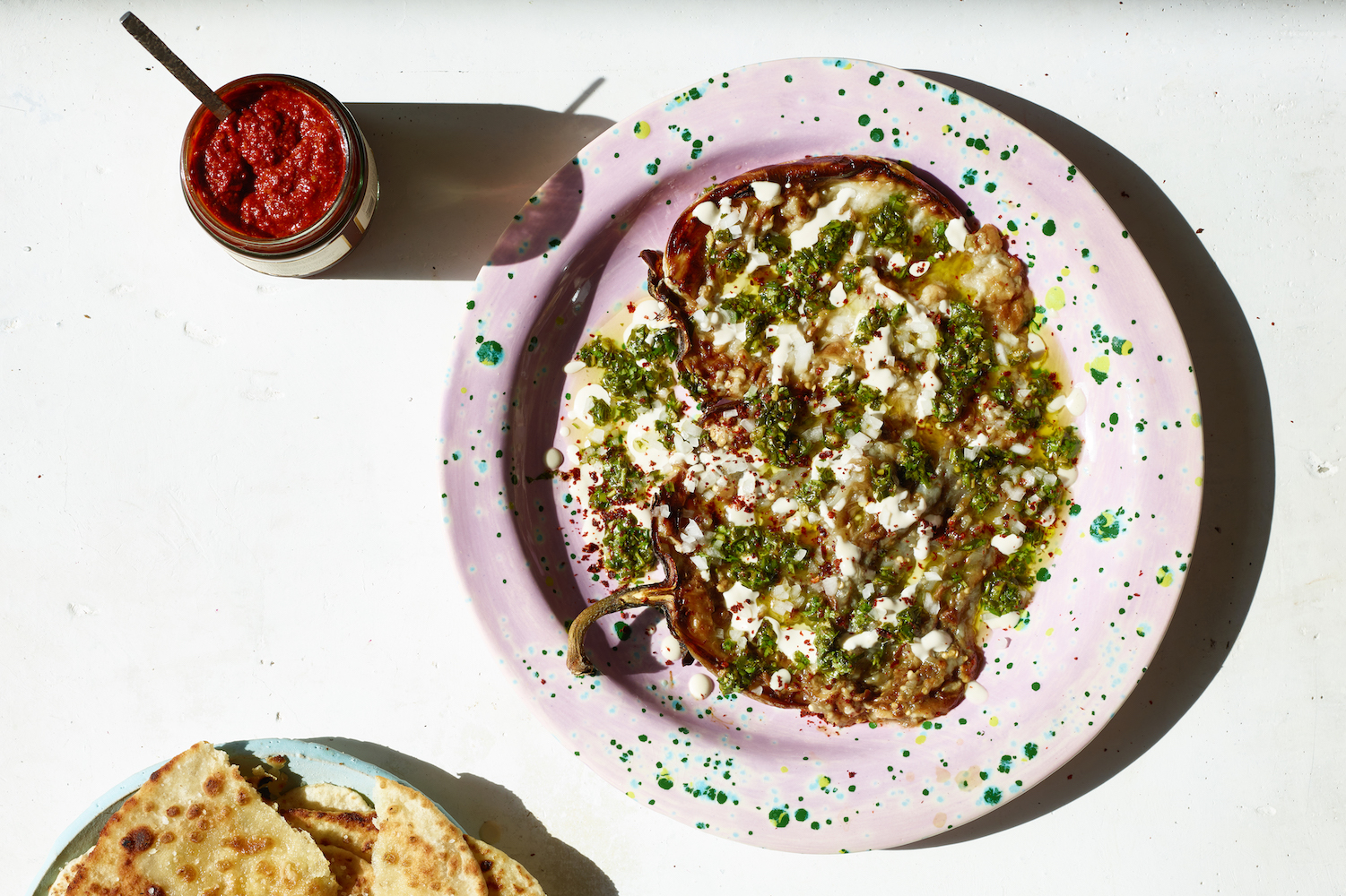 roasted eggplant dip on a speckled pink plate next to a plate of pita and jar of hot sauce