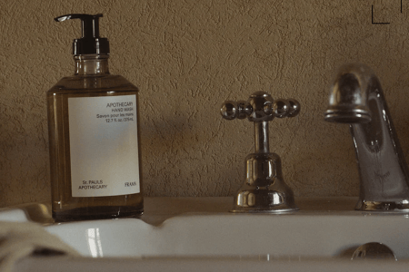 A brown glass bottle of "APOTHECARY HAND WASH" sits on a white sink next to a chrome faucet and tap.
