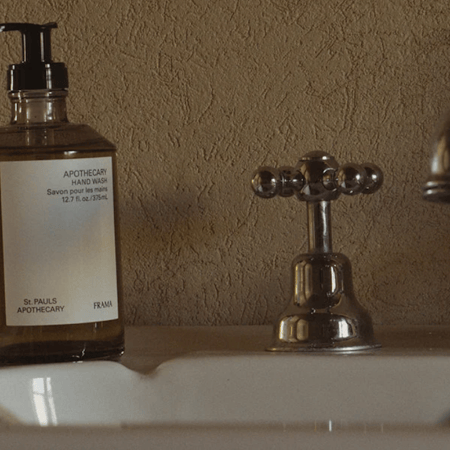 A brown glass bottle of "APOTHECARY HAND WASH" sits on a white sink next to a chrome faucet and tap.