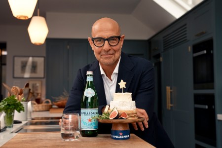 stanley tucci wearing a navy suit standing behind a wood-topped kitchen island with a bottle of S.Pellegrino and a tower of cheese