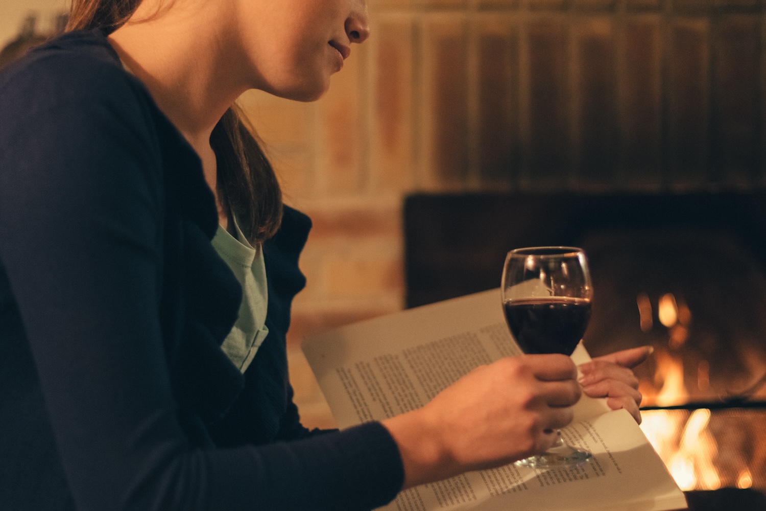 Woman near the fireplace reading a book with a glass of wine