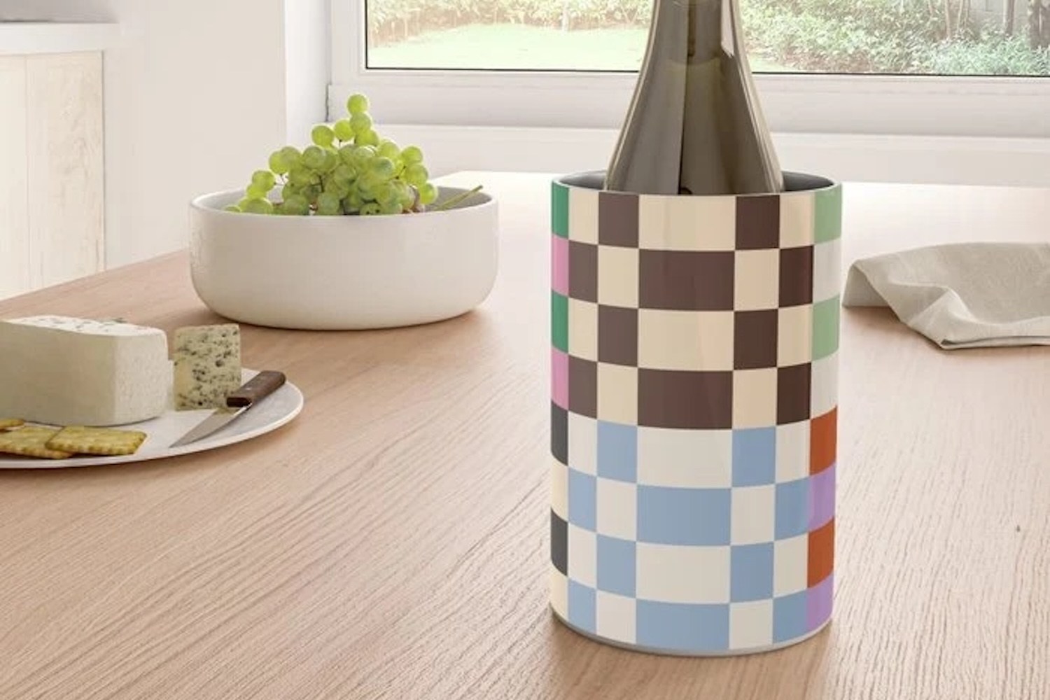 A wine bottle in a colorful checkered cooler on a wooden table with grapes, cheese, and crackers.