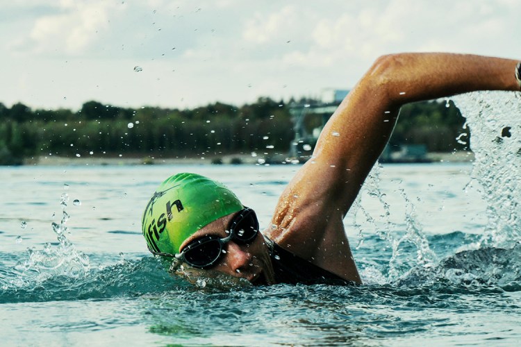 Adult swimmer wearing green swim cap and goggles doing freestyle stroke in open water, overlaid with bold green text reading "It’s Never Too Late."