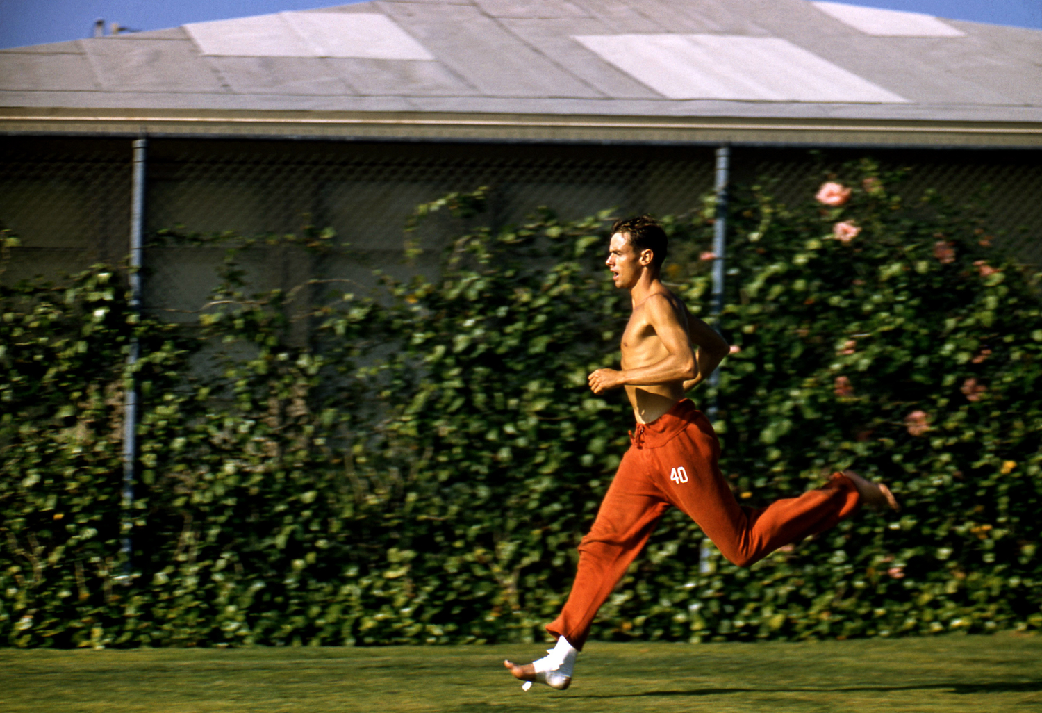 Shirtless male runner in red track pants sprinting on grass with gauze around his feet.