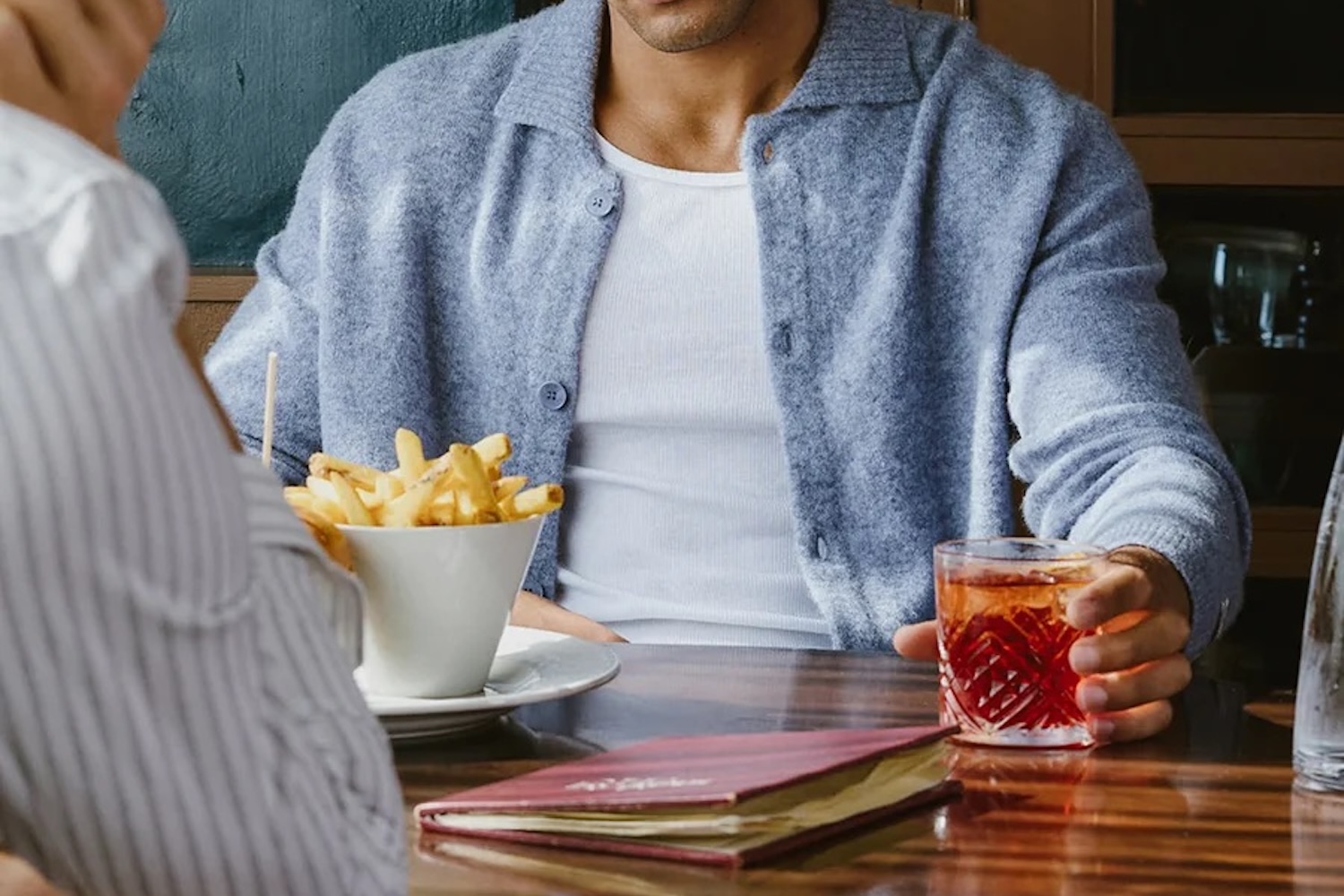 A person in a blue cardigan and white t-shirt holds a drink, with fries and a menu on the table.