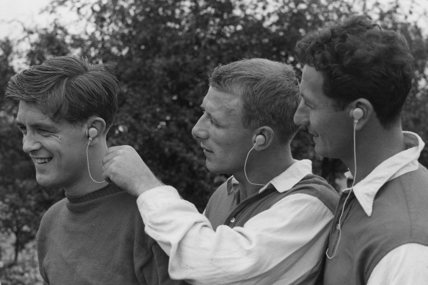 Three men wearing vintage-style wired headphones, smiling and interacting outdoors in a retro black-and-white photo.