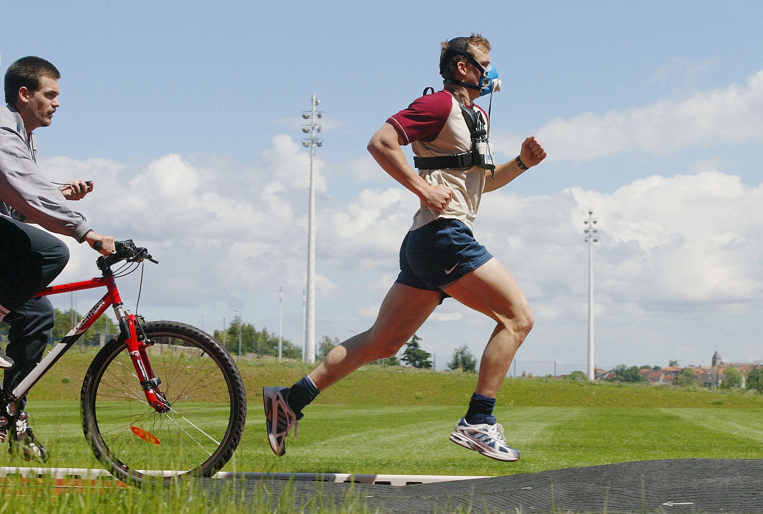 Man running with VO2 max mask on outdoor track next to coach on bike, symbolizing modern fitness optimization culture