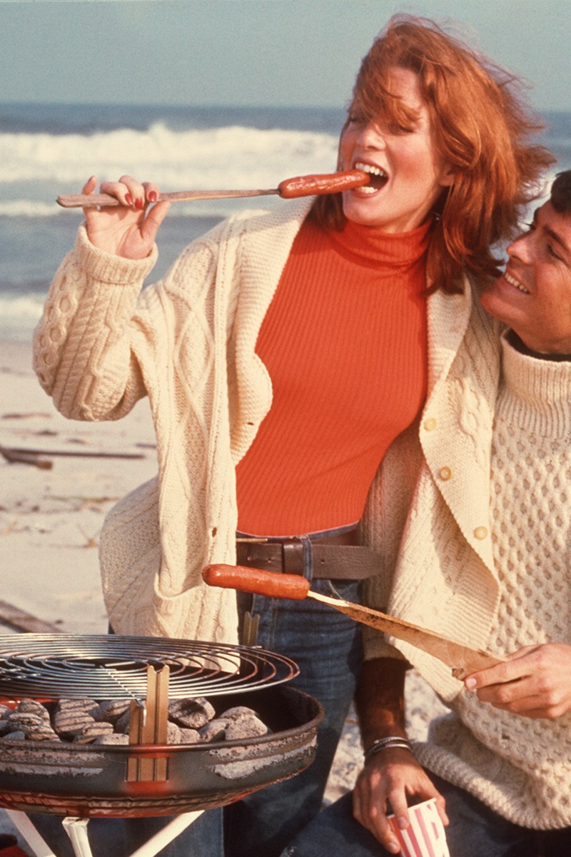 A man and woman enjoying a grilled hot dog on the beach.