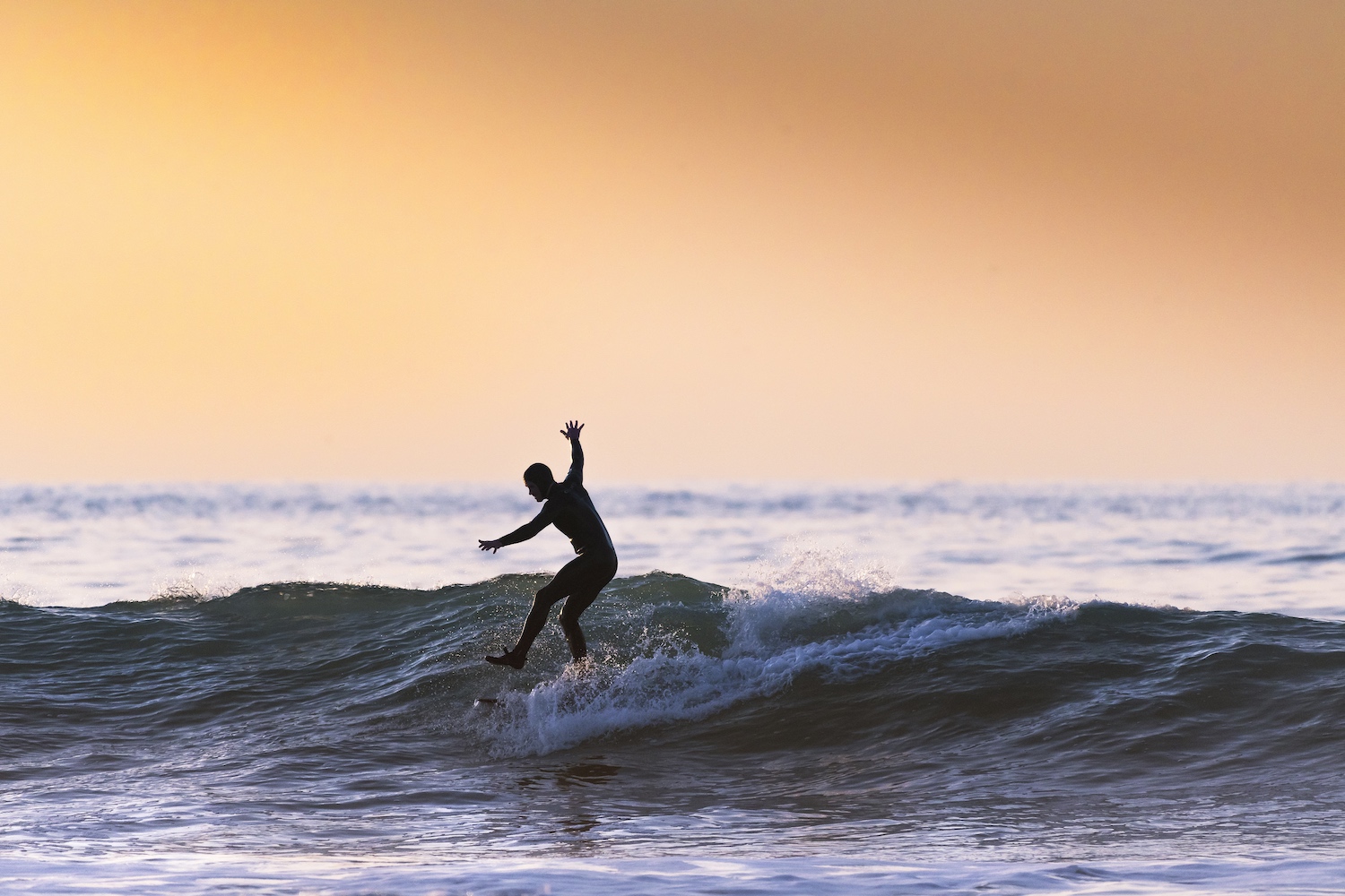 A surfer balancing on one leg while riding a wave at sunset.