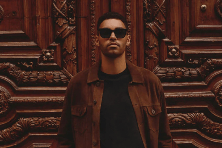 Man in sunglasses and a brown jacket standing in front of an ornate wooden door.