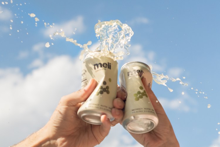 two people cryshing cans of meli beer in front of a blue sky