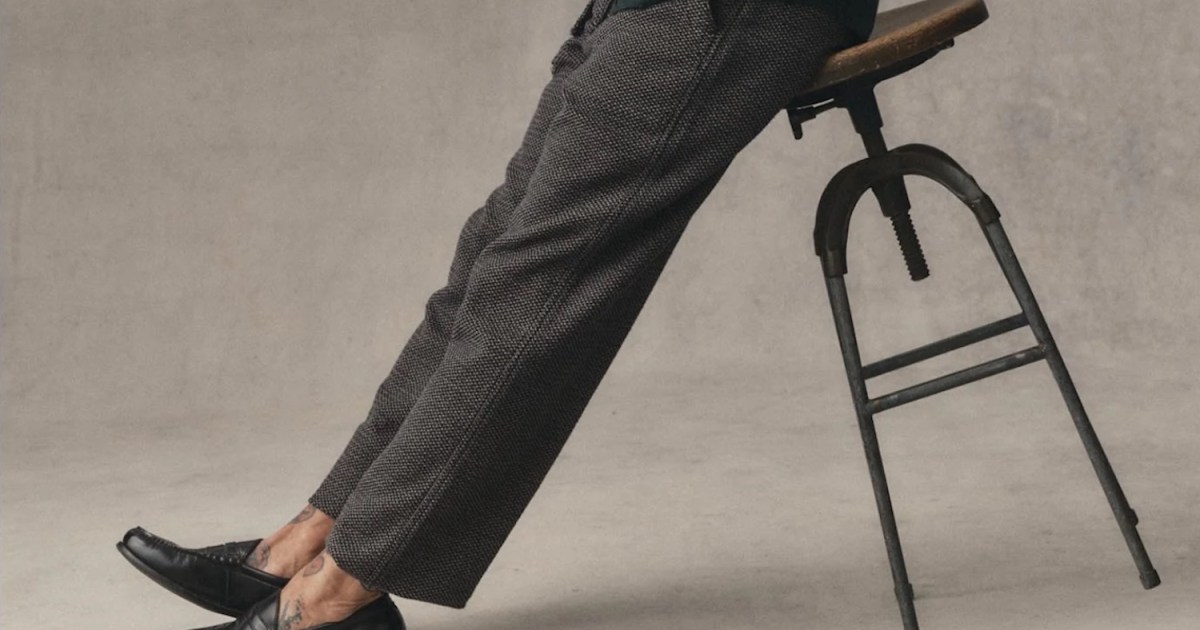Person in patterned trousers and black loafers leaning on a wooden stool, revealing ankle tattoos.