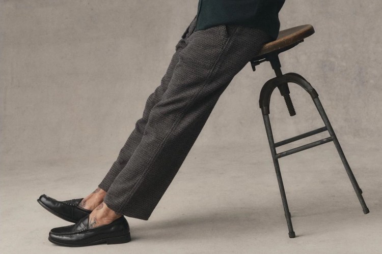 Person in patterned trousers and black loafers leaning on a wooden stool, revealing ankle tattoos.