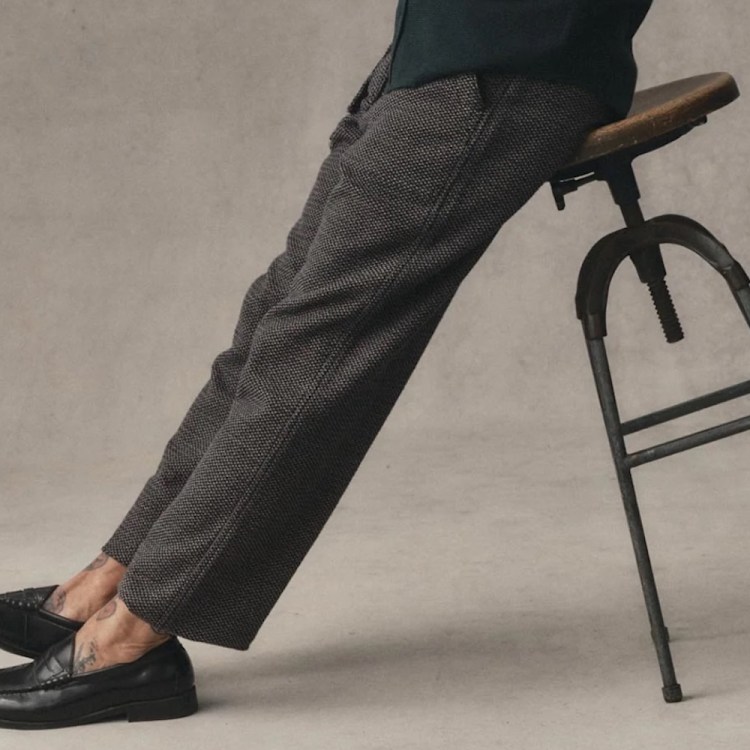 Person in patterned trousers and black loafers leaning on a wooden stool, revealing ankle tattoos.