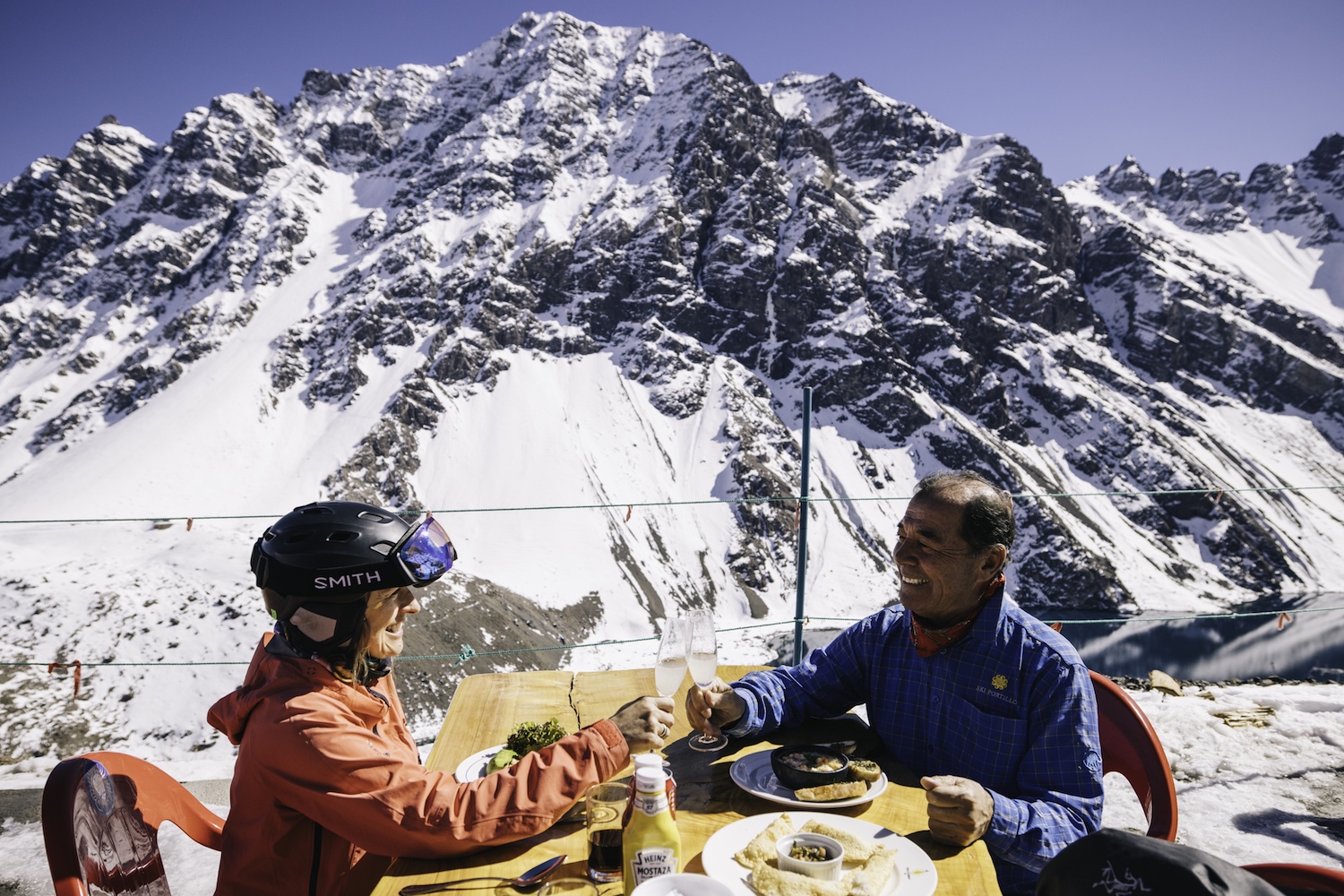 Two skiers share a drink at a rooftop bar with mountains in the background.