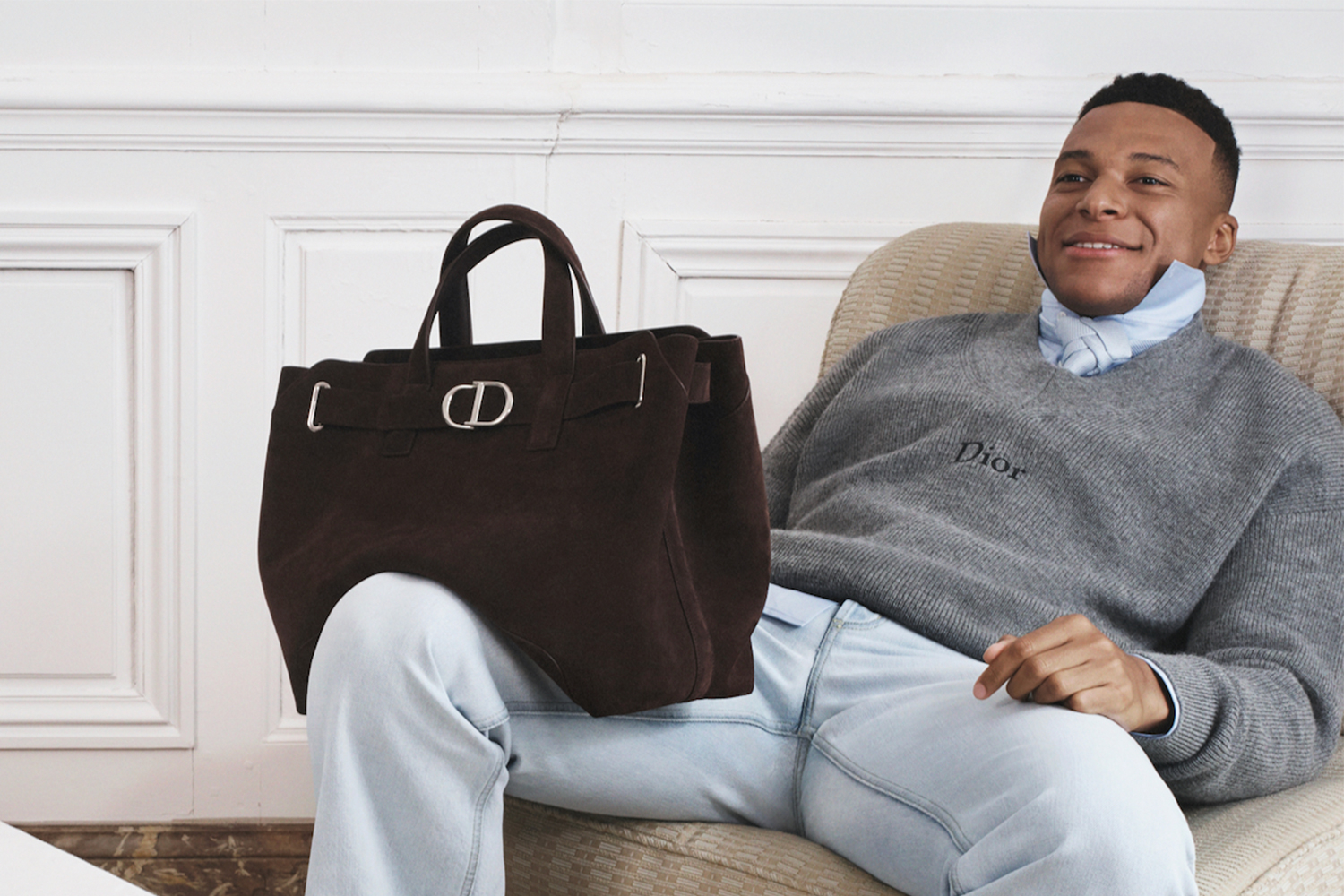 Soccer player Kylian Mbappé sitting on a chair with a brown suede Dior bag