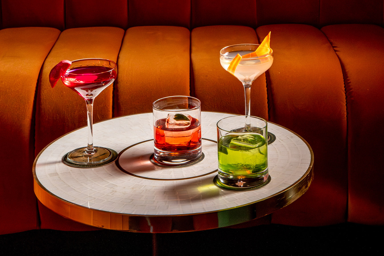 A small table topped with four cocktails, including Gin & Tonics and Martinis, in front of an orange banquette