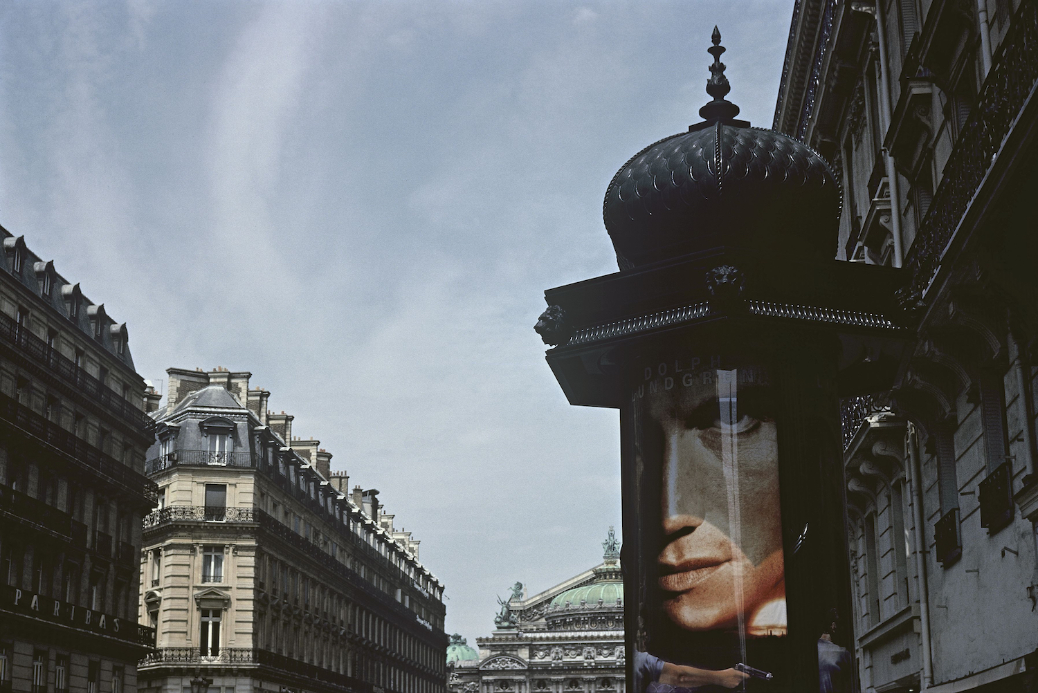 A view of a Paris street, with a Morris column in the foreground displaying an advertising poster