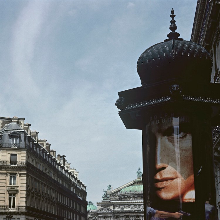 A view of a Paris street, with a Morris column in the foreground displaying an advertising poster