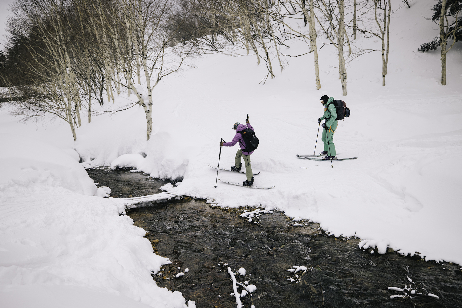 Two skiers cross a little stream in the snow.