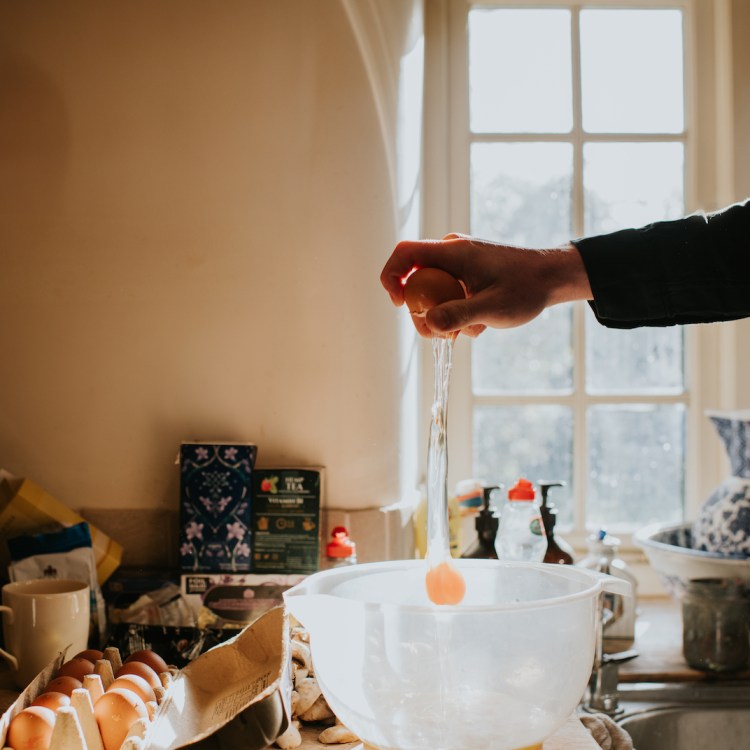 A man breaks an egg into a mixing bowl from a height, using one hand.