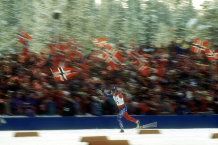 A Nordic skier lunges for the finish line, with Norwegian fans in the stands behind him.