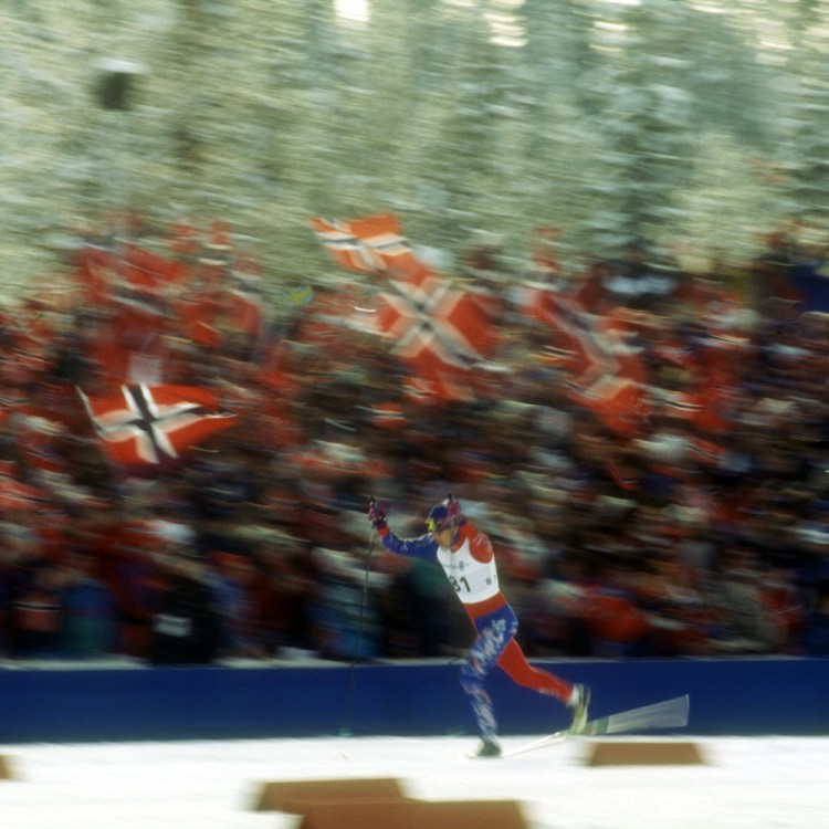 A Nordic skier lunges for the finish line, with Norwegian fans in the stands behind him.