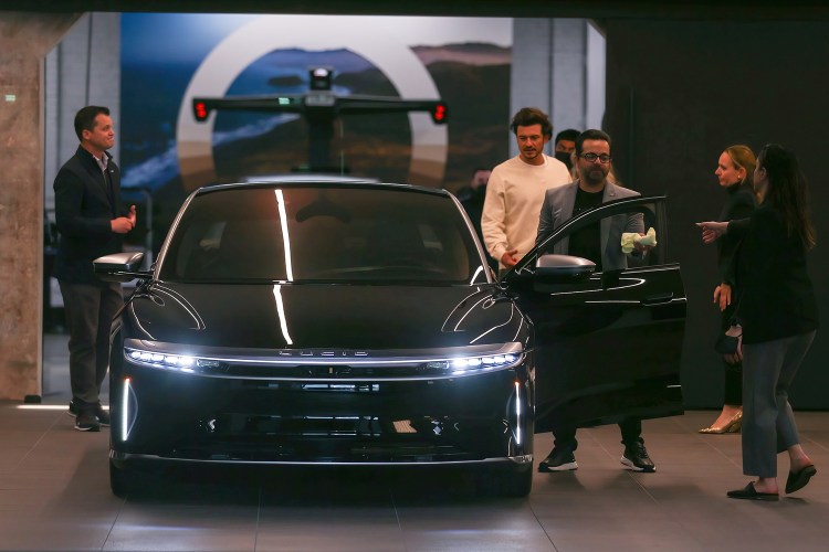Orlando Bloom in a white sweatshirt picking up a luxury EV called the Lucid Air