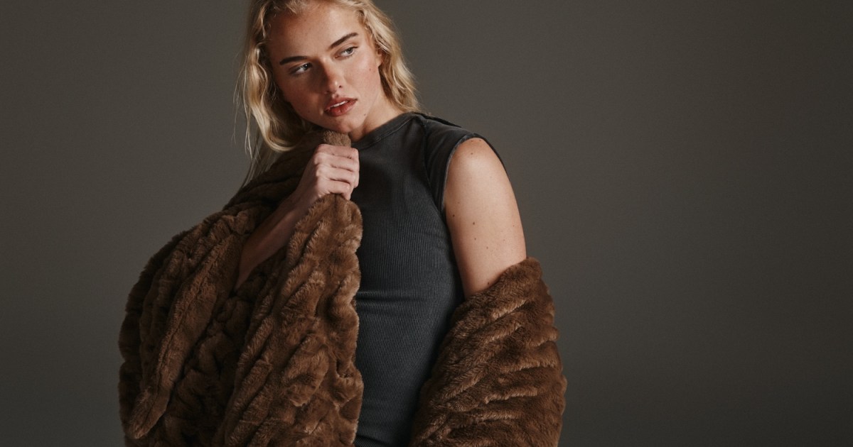 a blond woman standing against a gray background wrapped in a plush brown lola blanket