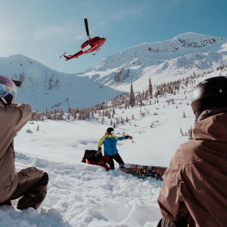 A trio of people hanging out in snow as helicopter takes off.