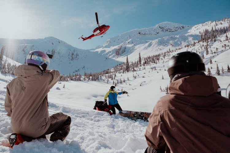 A trio of people hanging out in snow as helicopter takes off.