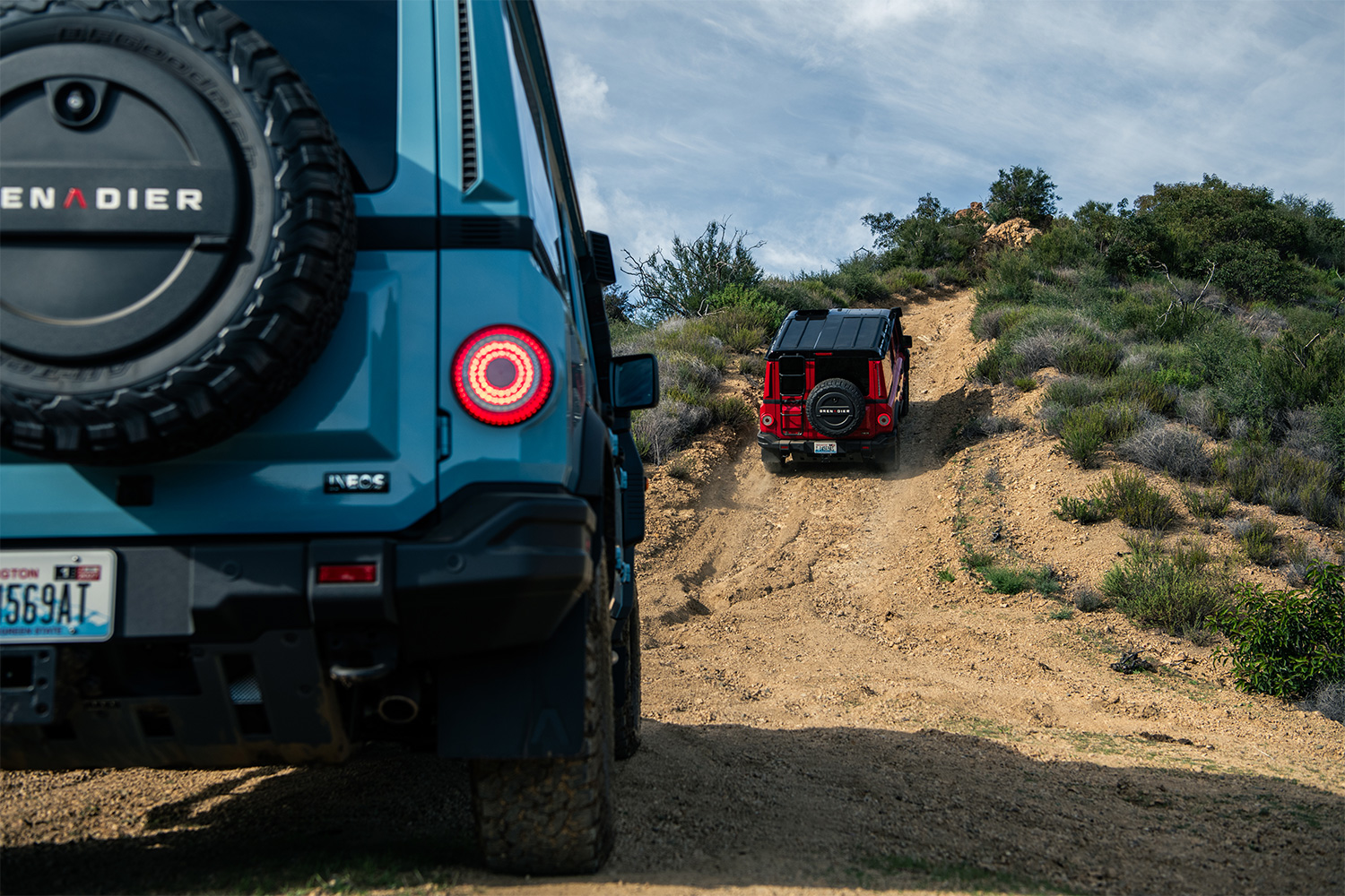 A blue Ineos Grenadier SUV following a red version of the off-road vehicle up a dirt hill
