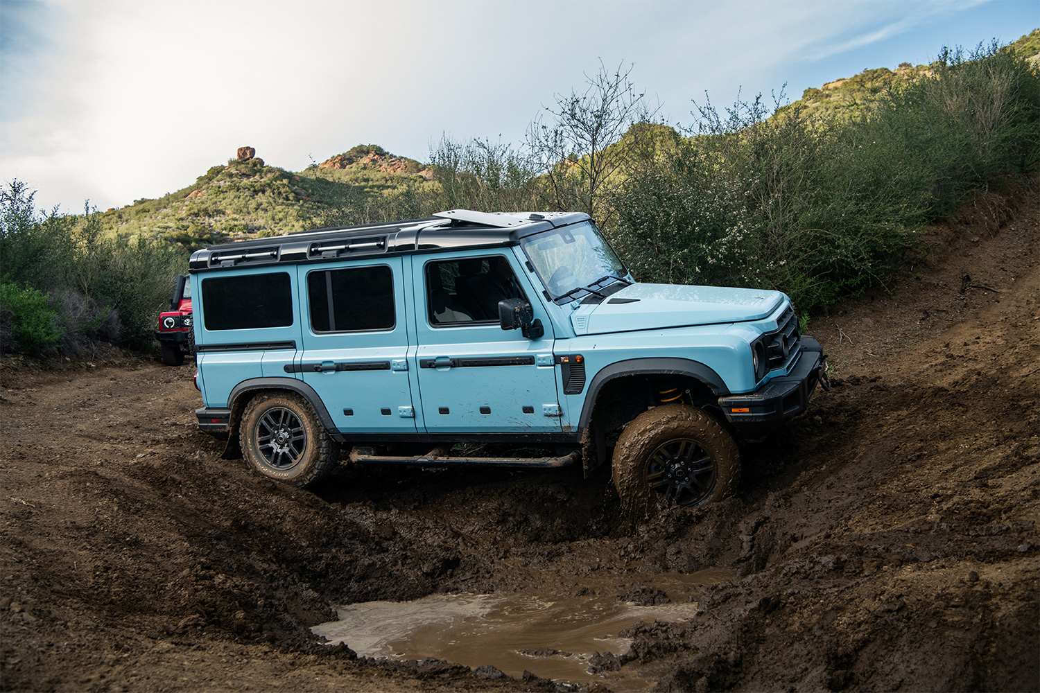 A blue 2026 Ineos Grenadier SUV driving through a deep mud pit