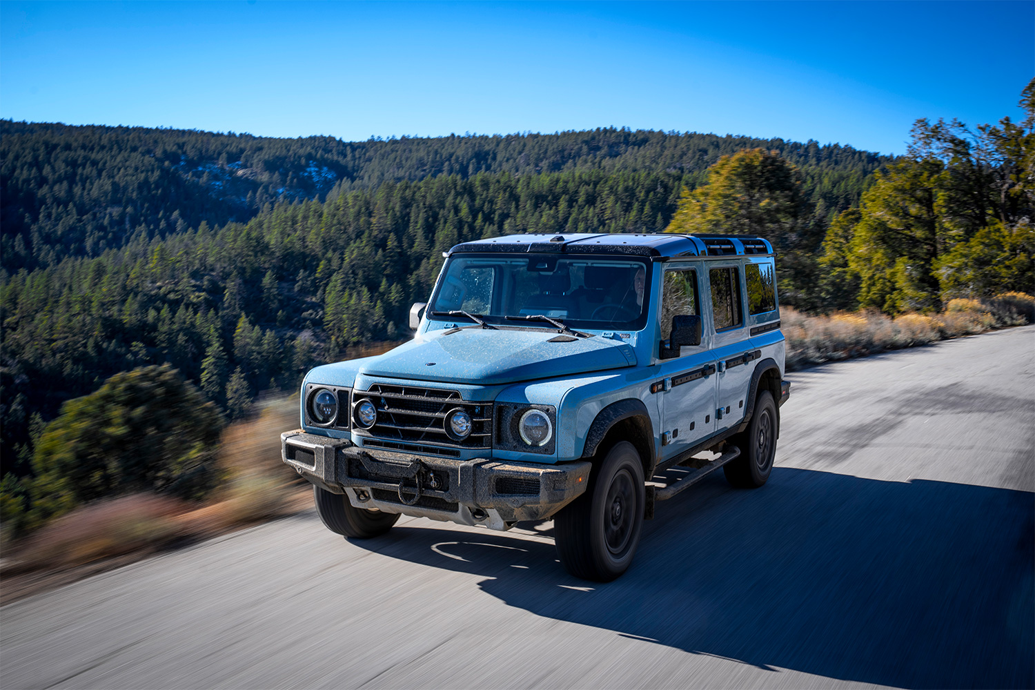 A blue 2026 Ineos Grenadier Station Wagon (or SUV) driving along a paved road next to a forest
