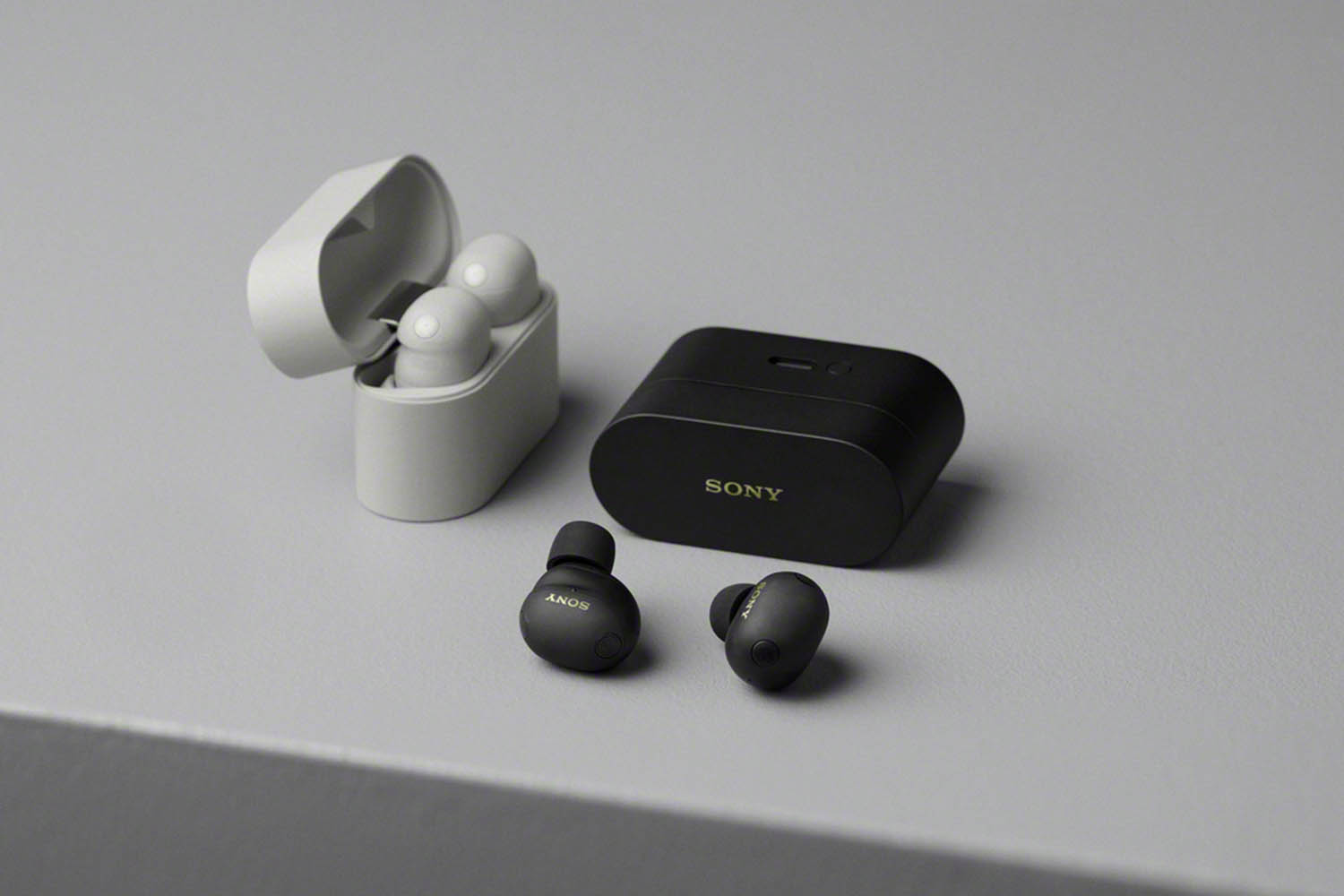 A pair of black earbuds on a table next to a case and a pair of white earbuds in a case