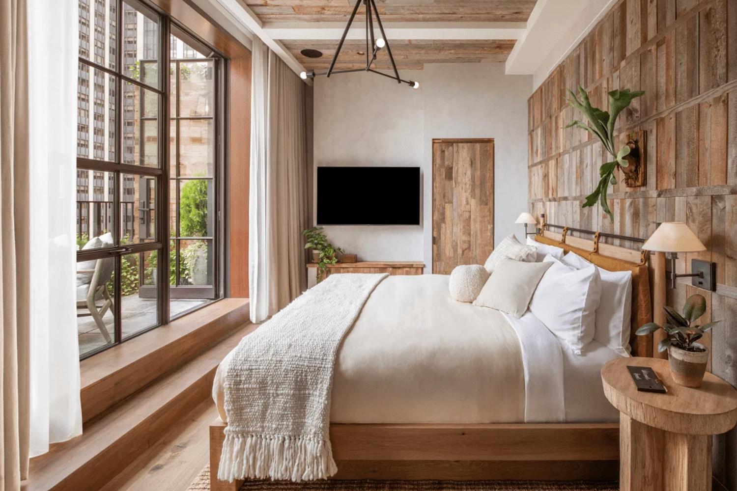 Guestroom suite at 1 Hotel Central Park featuring nature-inspired design with reclaimed wood finishes, neutral tones, abundant greenery and large windows offering views of Midtown Manhattan.