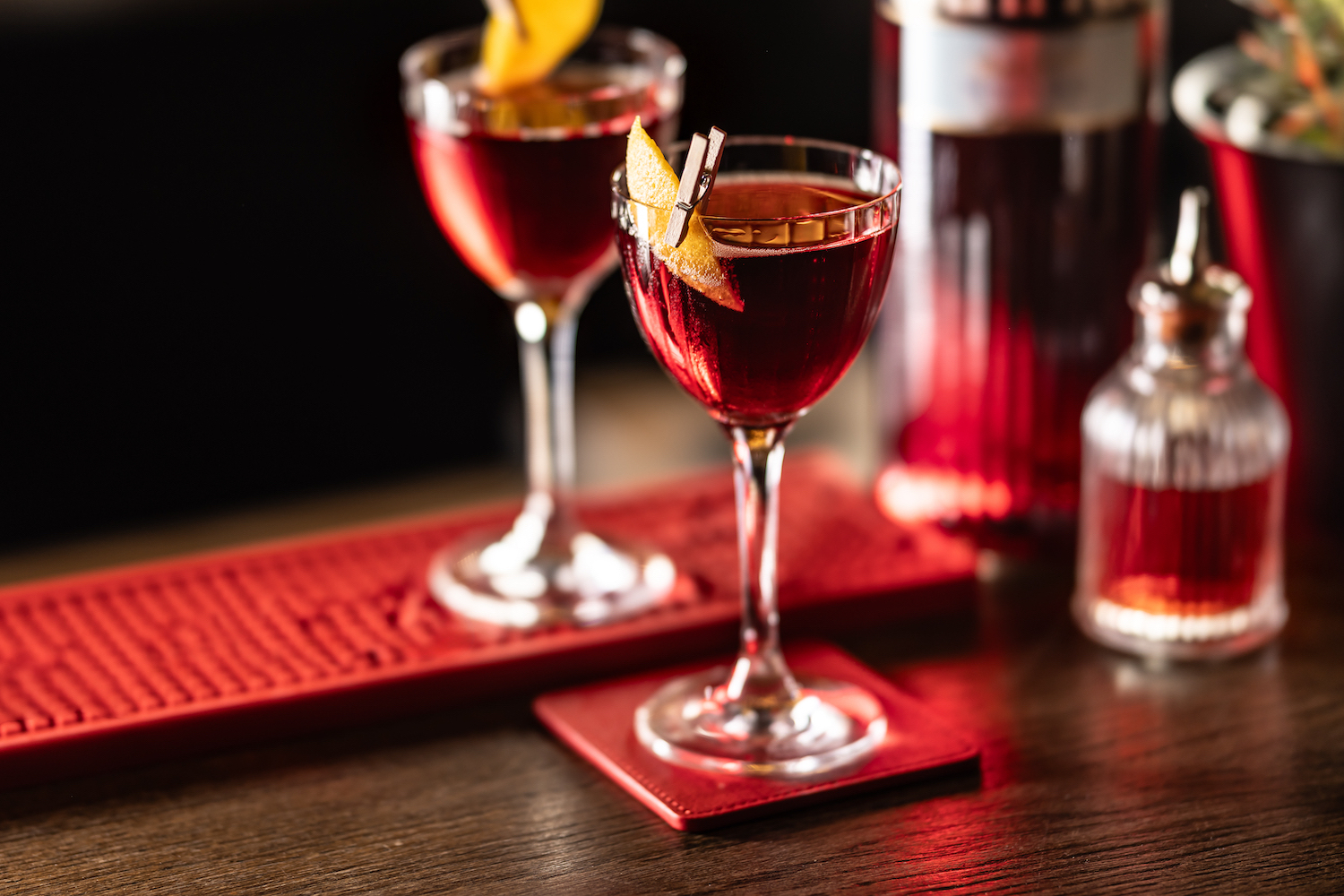 Two Negroni cocktails in stemmed glasses, each garnished with a twist of orange peel, set on the bar on a red bar mat and red coasters