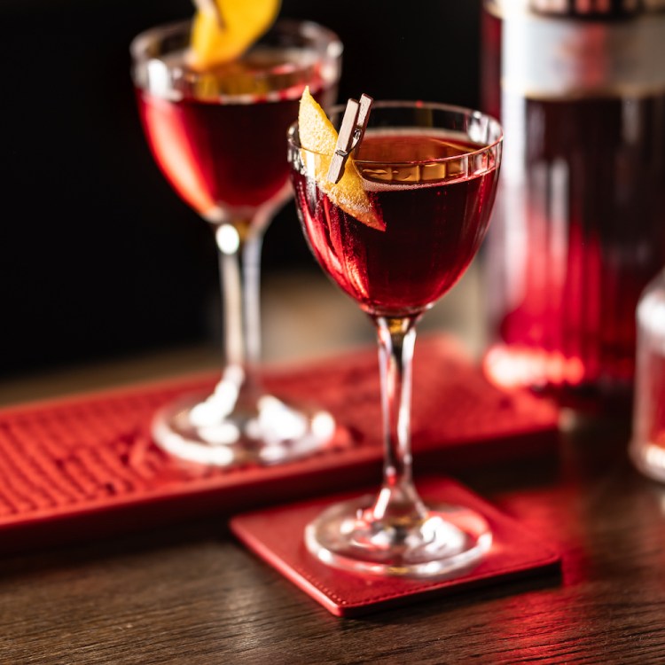 Two Negroni cocktails in stemmed glasses, each garnished with a twist of orange peel, set on the bar on a red bar mat and red coasters