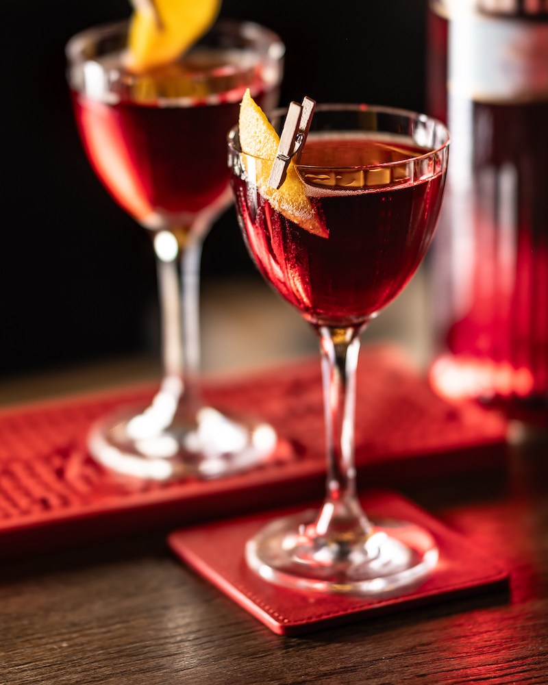 Two Negroni cocktails in stemmed glasses, each garnished with a twist of orange peel, set on the bar on a red bar mat and red coasters