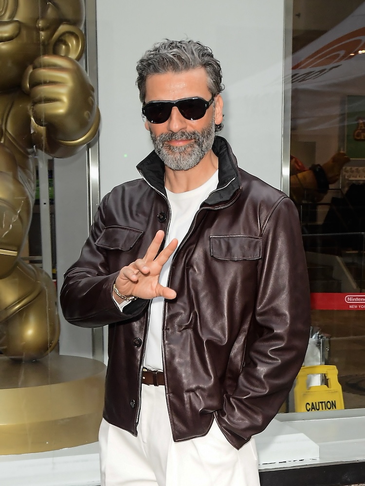A man in sunglasses and a leather jacket waves with one hand