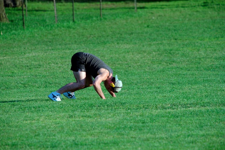 A man working out on all fours in a green field with a hat and headphones on